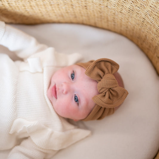 Newborn baby wearing a brown waffle top knot headband with a bow, lying on a white blanket.