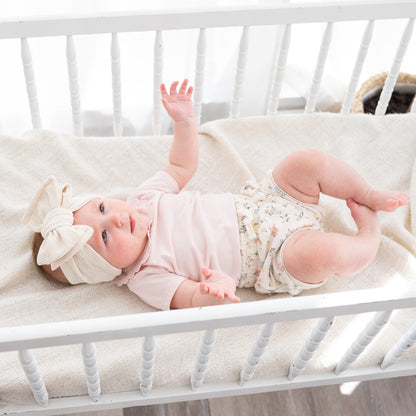 Baby lying in a crib wearing a pink outfit and beige waffle oversized bow headband.