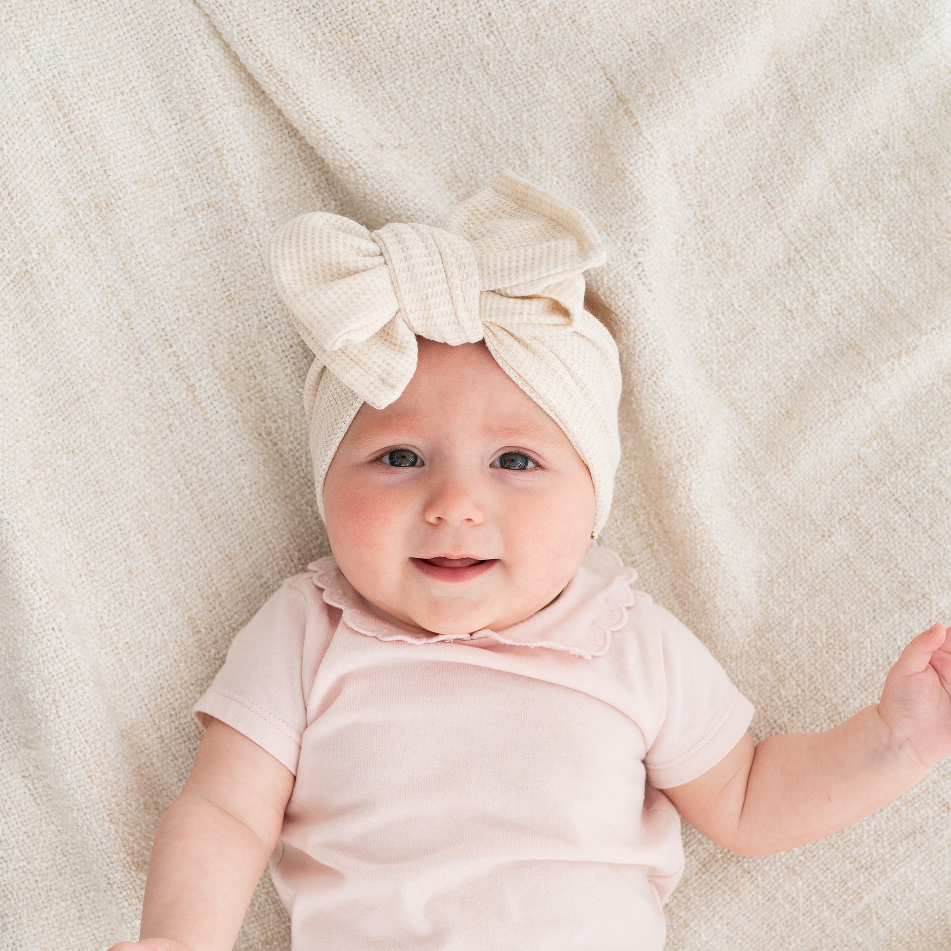 Baby wearing a beige waffle bow headband and pink outfit on a soft beige blanket