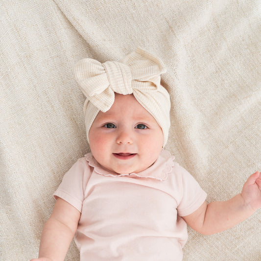 Baby wearing a beige waffle bow headband and pink outfit on a soft beige blanket