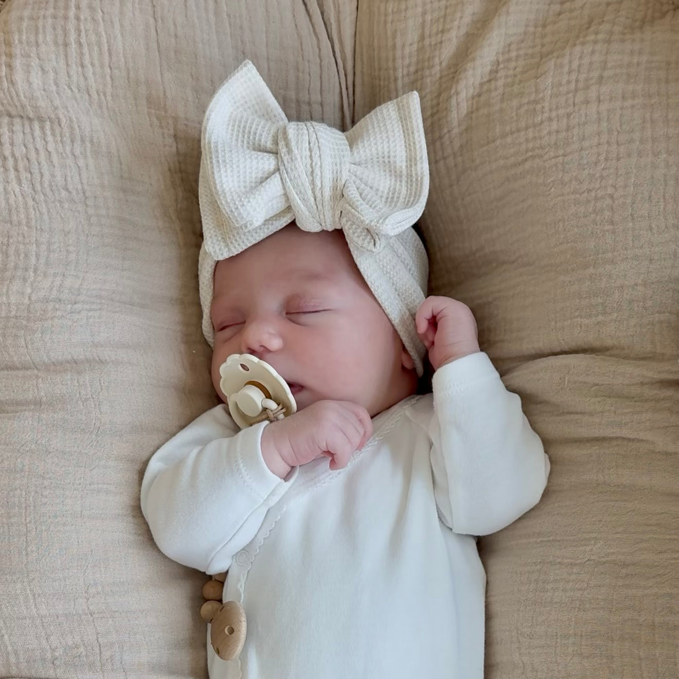 Newborn baby wearing a white outfit and beige waffle headband with a bow, lying on a beige blanket.