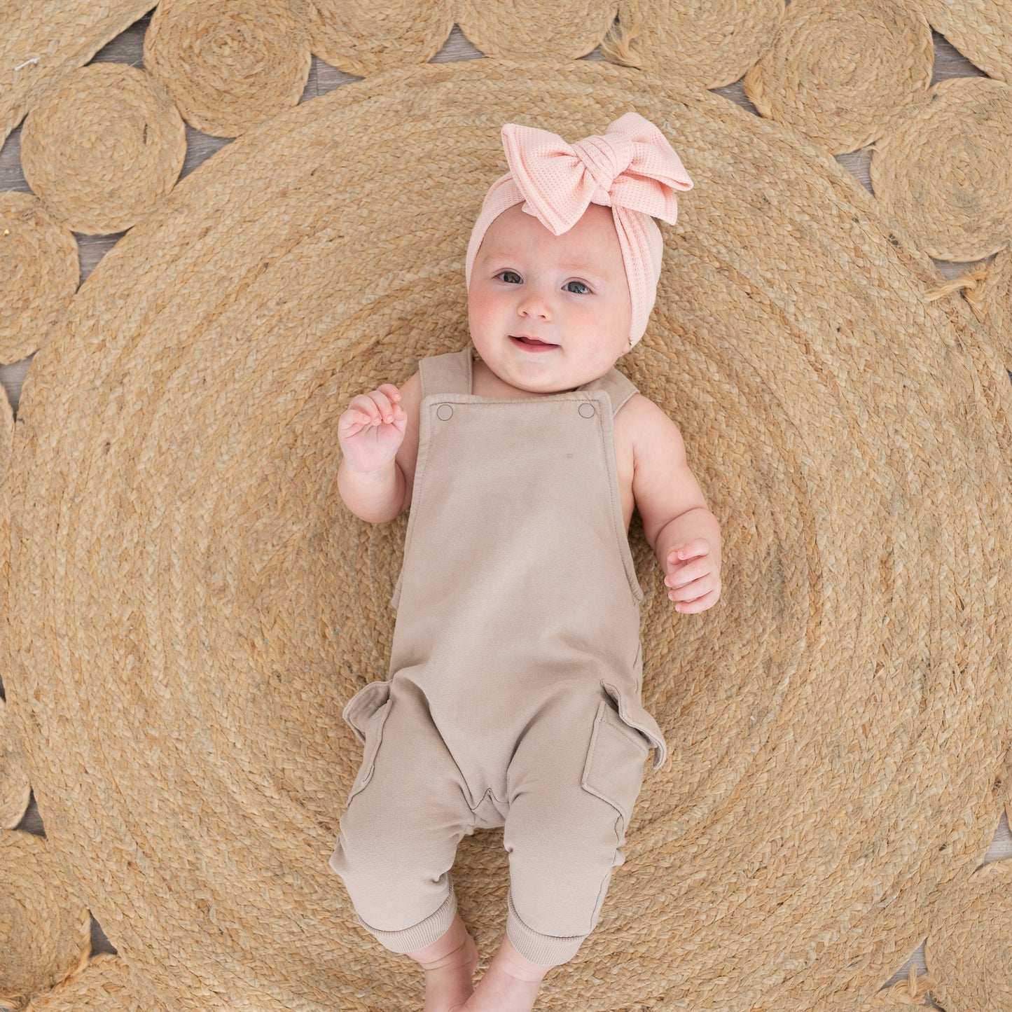 Baby in beige overalls and pink waffle oversized bow headband lying on a woven mat