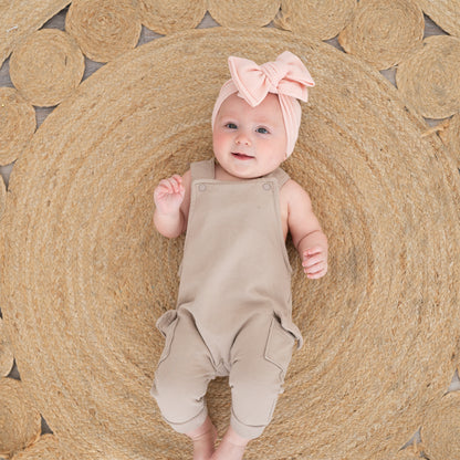 Baby in beige overalls and pink waffle oversized bow headband lying on a woven mat