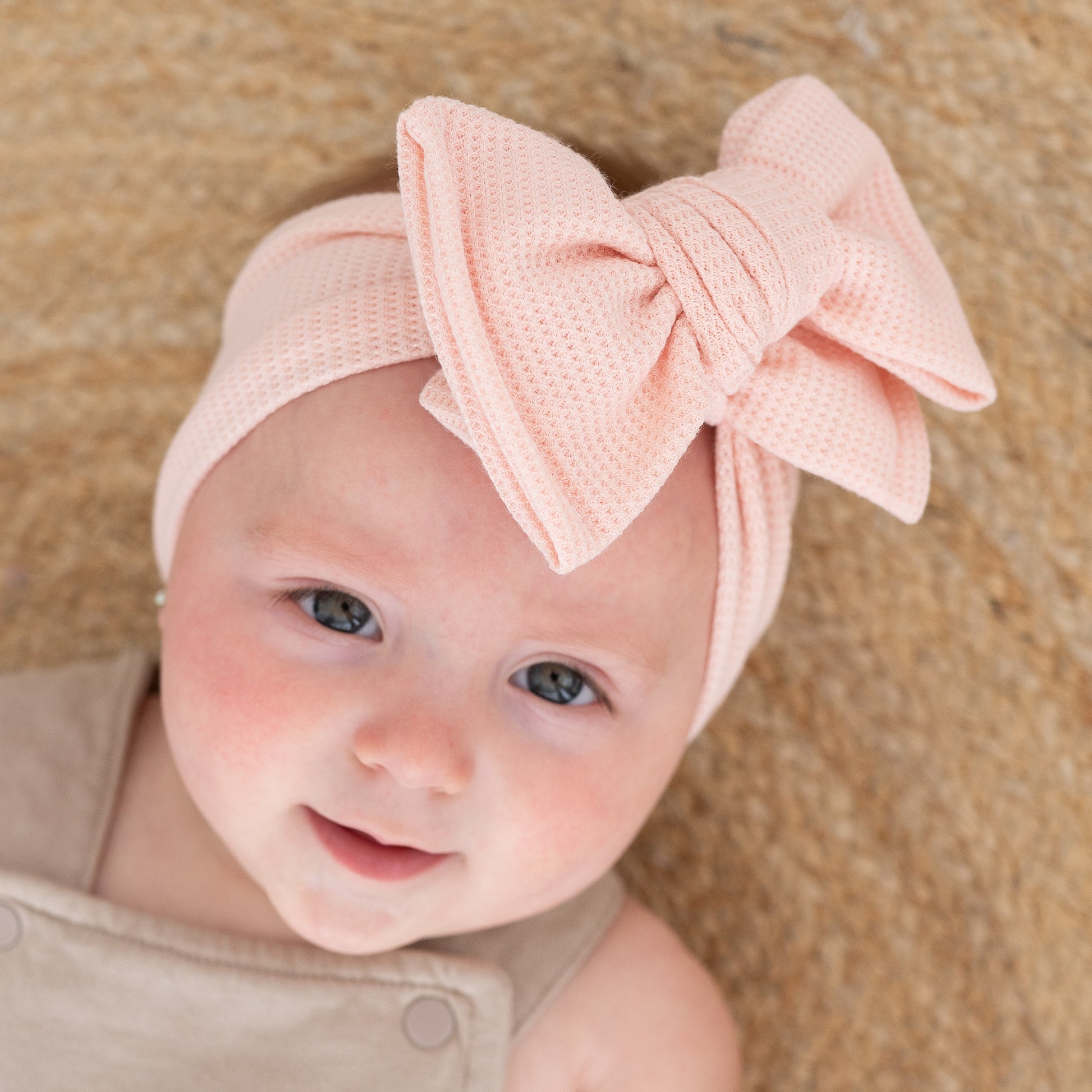 Closeup of a baby girl's face, wearing a pink waffle headband with a large bow on a beige background
