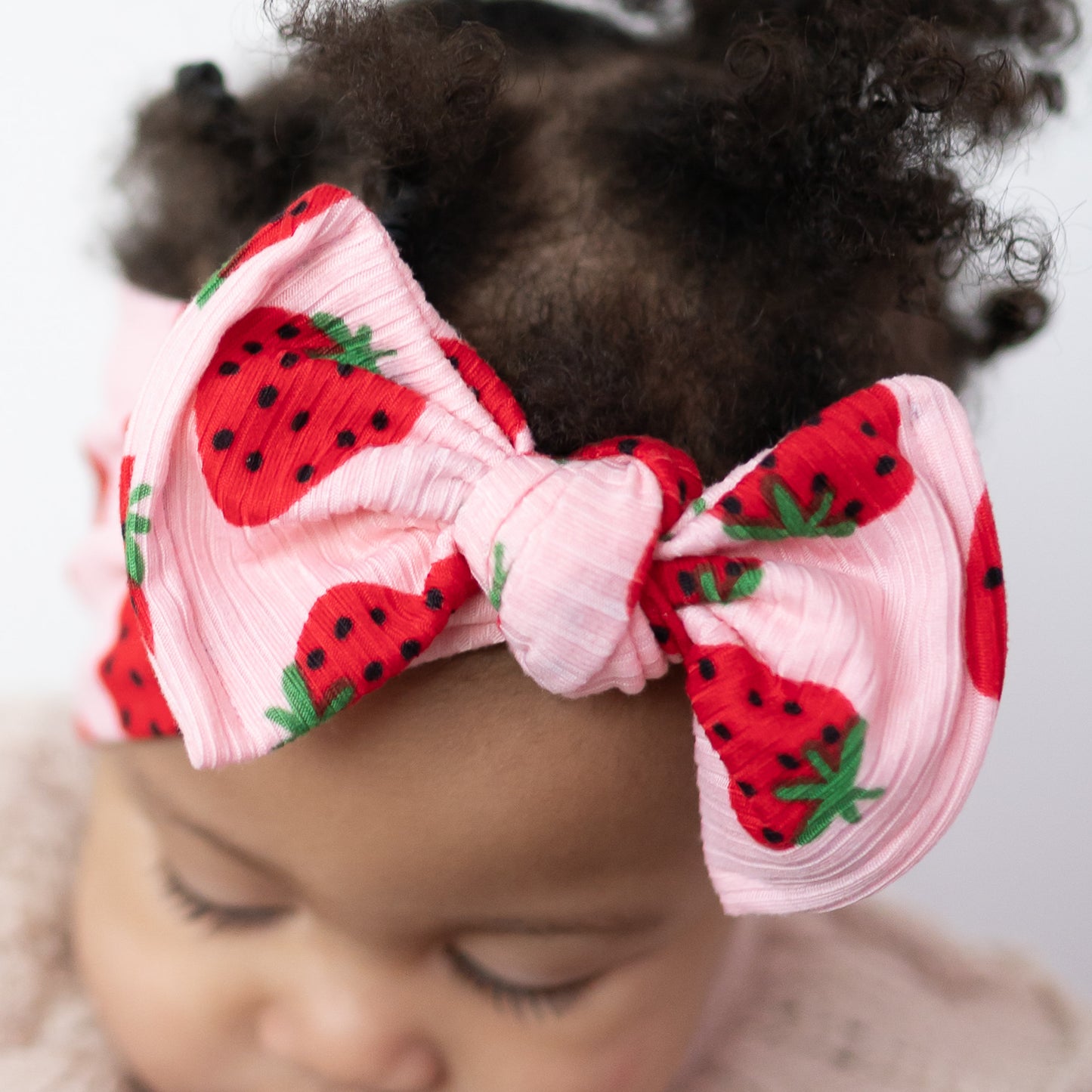 Detail view of baby girl with curly hair wearing a headband with strawberry designs on a plain background