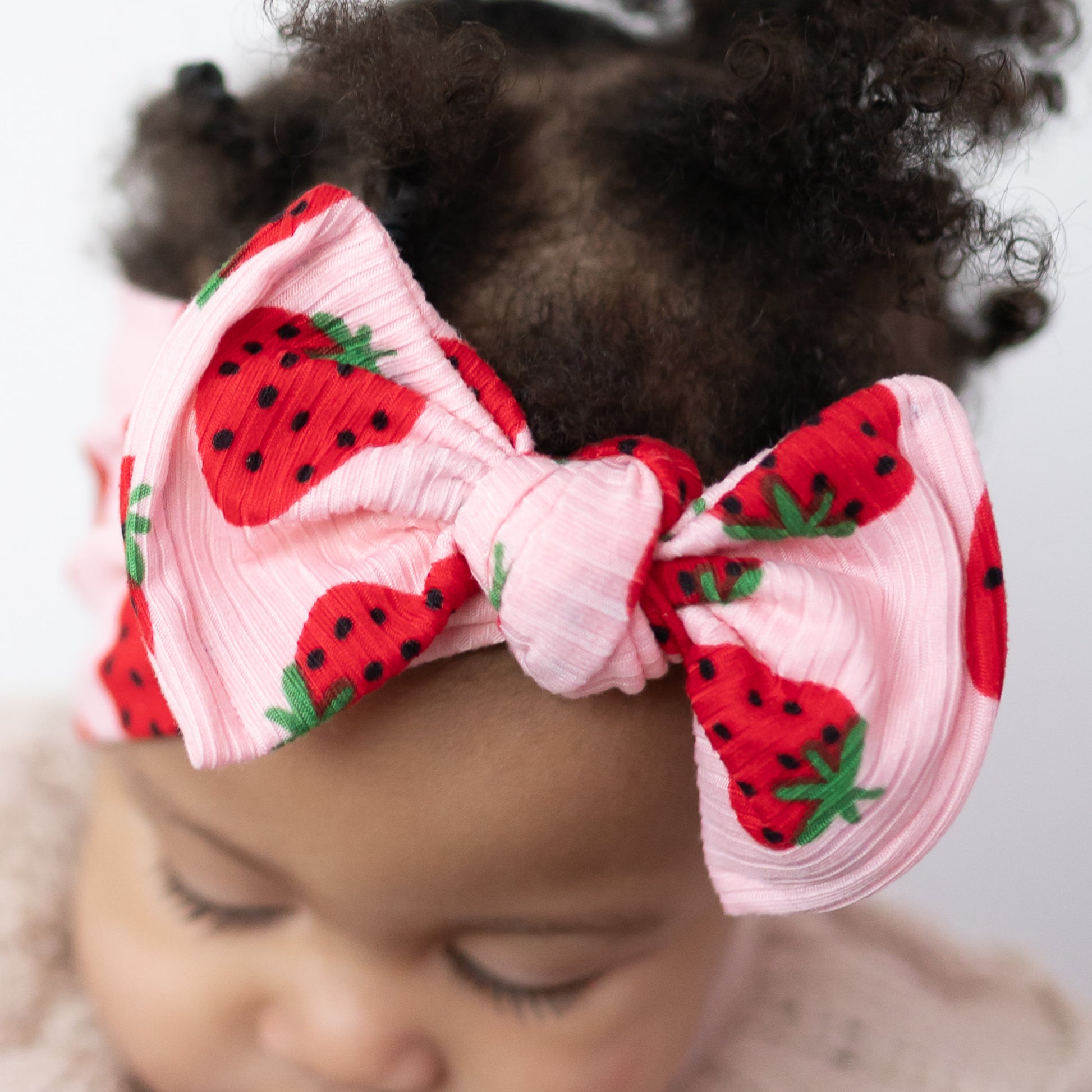 Detail view of baby girl with curly hair wearing a headband with strawberry designs on a plain background