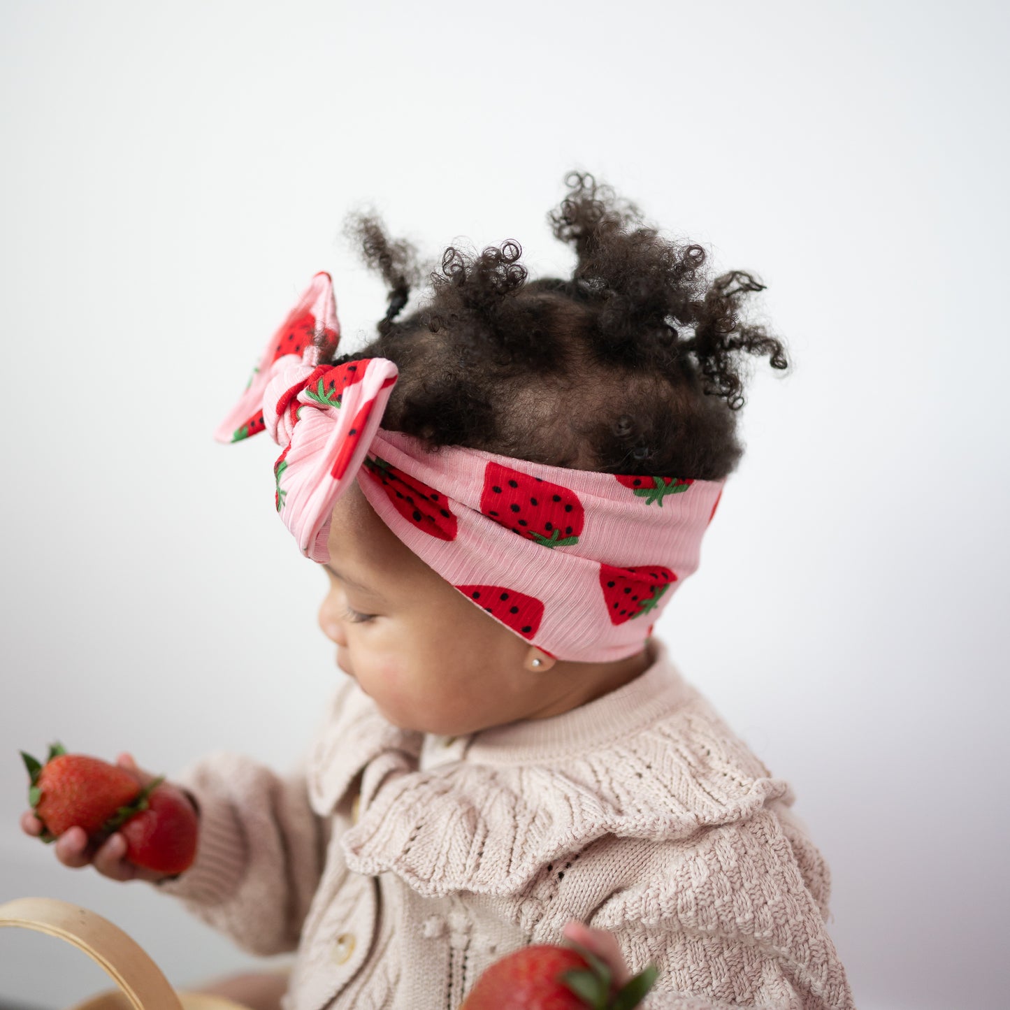 Child wearing a pink headband with red strawberry pattern, holding strawberries.