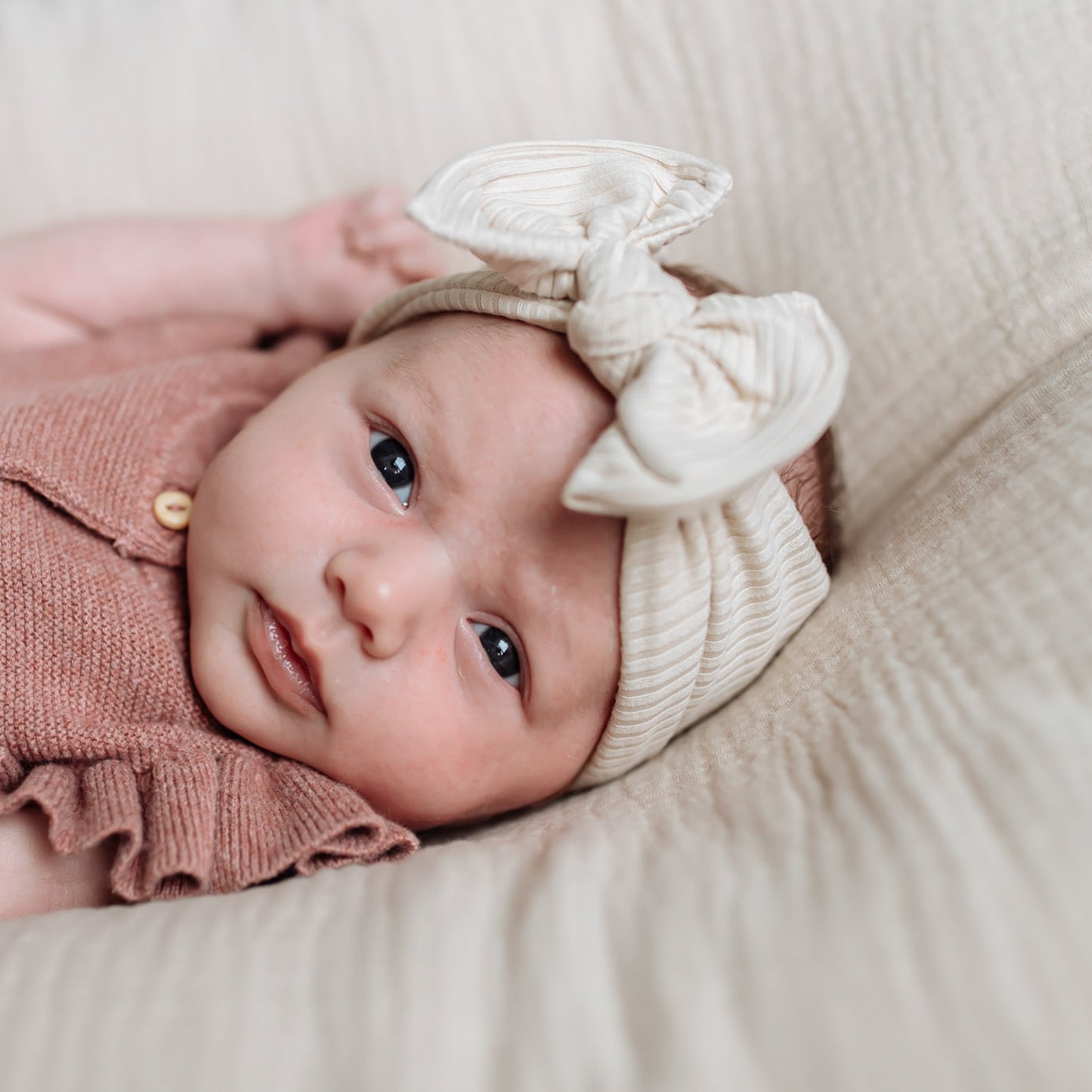 Newborn baby girl wearing a pink outfit and beige ribbed top knot headband with a bow, lying on a soft surface.