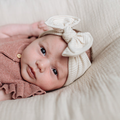 Newborn baby girl wearing a pink outfit and beige ribbed top knot headband with a bow, lying on a soft surface.