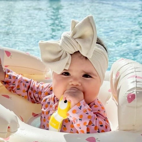 Baby with a large beige waffle bow headband sitting in a float in a pool