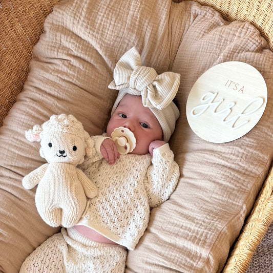 Newborn baby girl in a cream crochet outfit with a bow, holding a stuffed lamb and pacifier, lying on beige fabric with 'It's a Girl' sign.