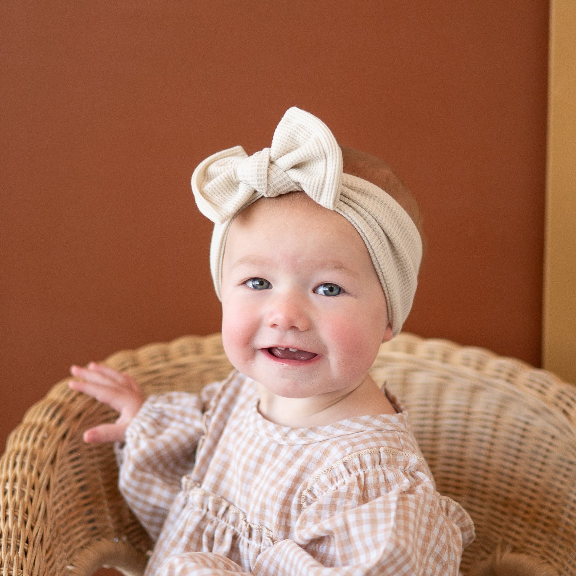 Baby wearing a beige headband with a bow, sitting in a wicker chair against a brown background