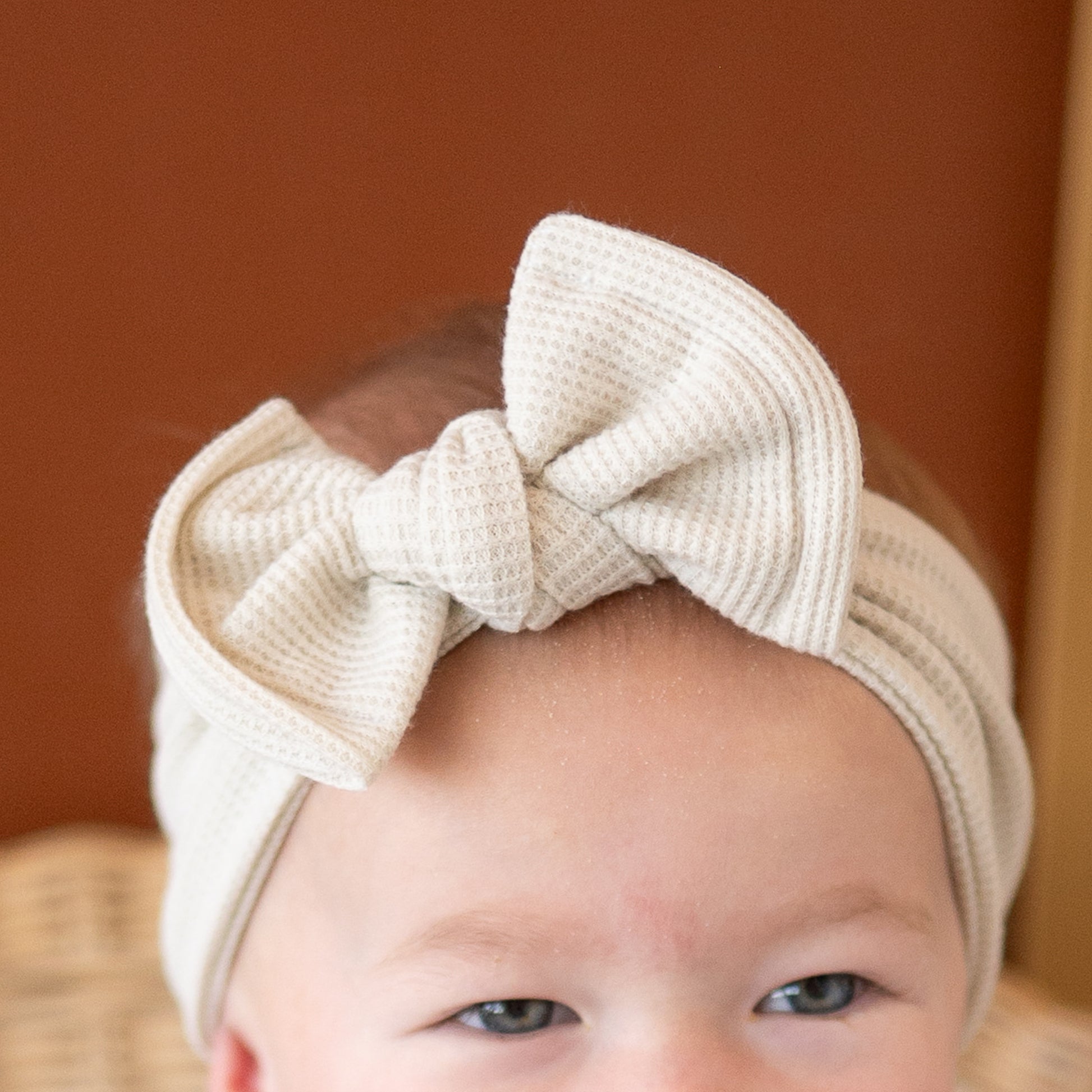 Close up view of a baby wearing a beige headband with a bow against a brown background