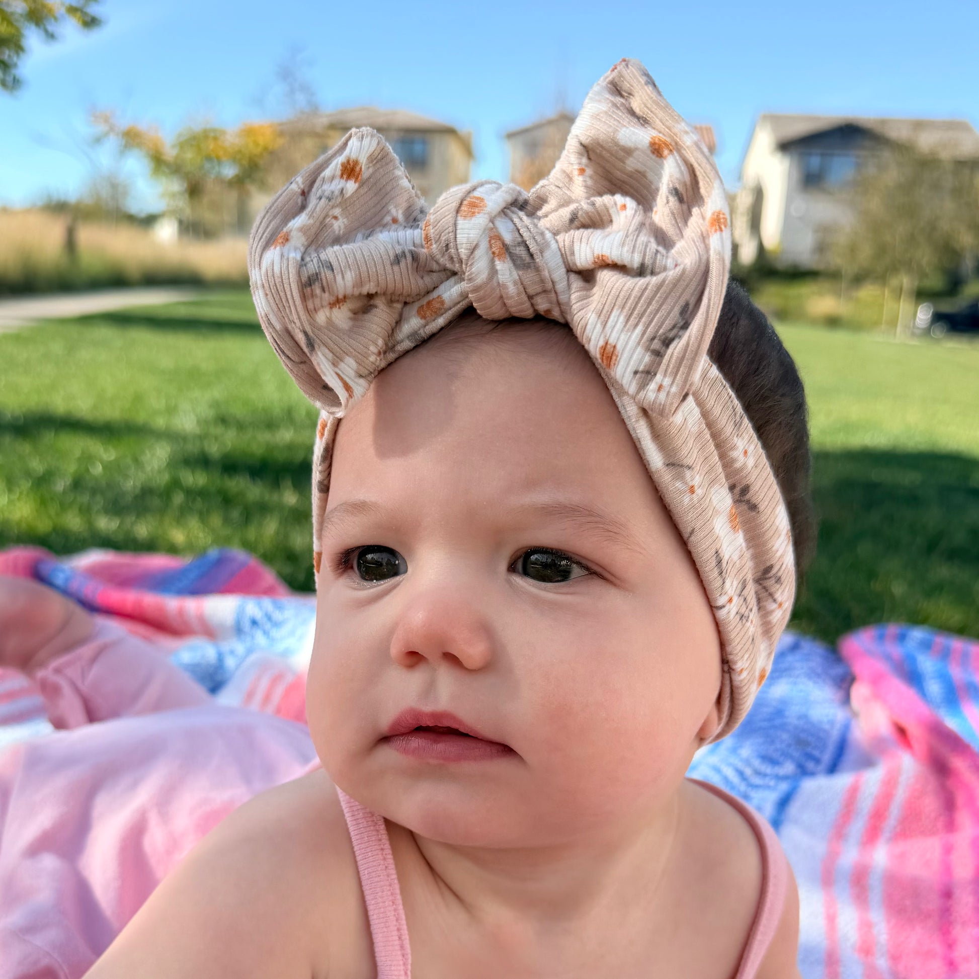 Baby with a large ribbed top knot bow headband sitting on a colorful blanket outdoors.