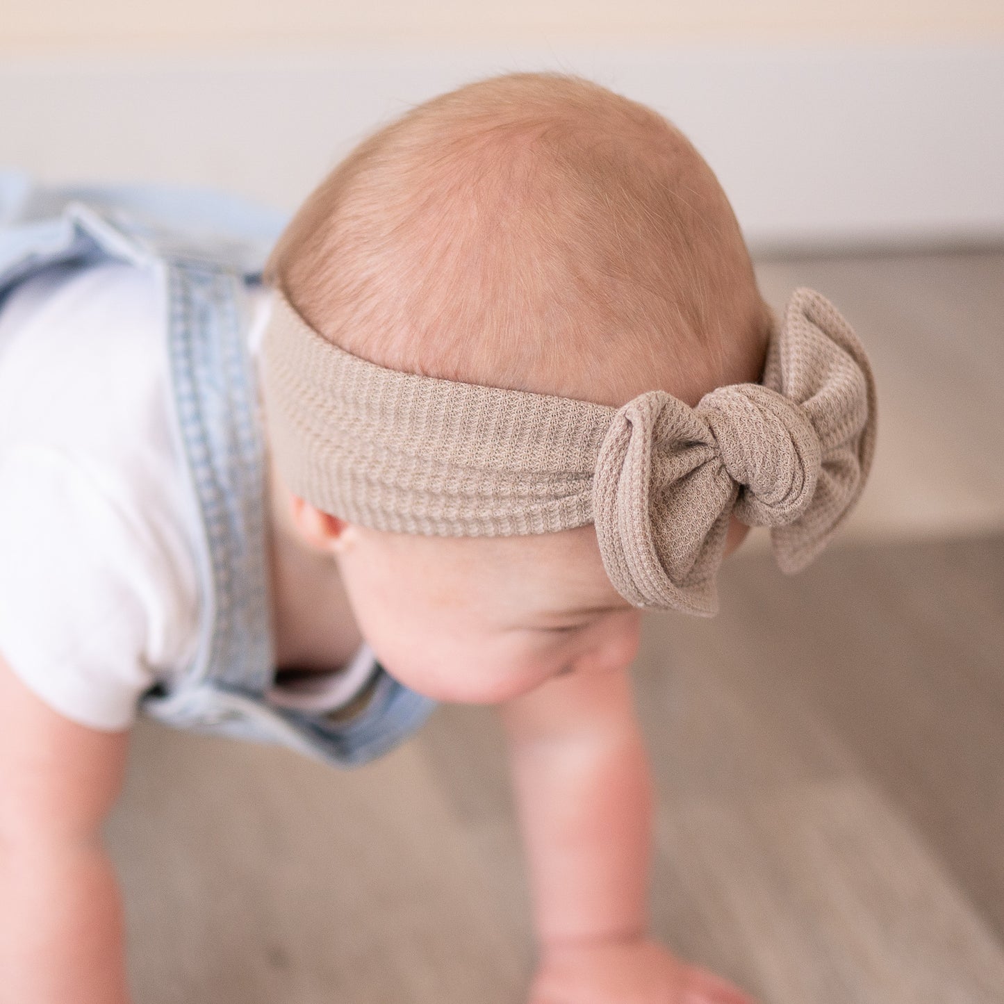 Side view of a baby wearing a taupe headband with a bow, looking down.