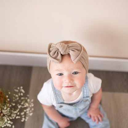 Baby wearing a taupe headband with a large bow, sitting on a wooden floor.