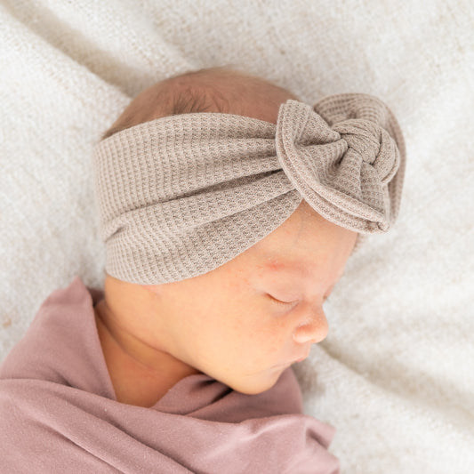 Newborn baby wearing a taupe top knot headband on a soft white blanket.