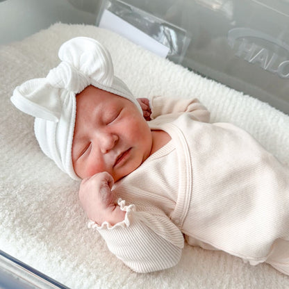 Newborn baby girl in a hospital cot wearing a white ribbed top knot bow.