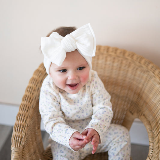 Baby wearing a white oversized bow bow headband sitting on a wicker chair.