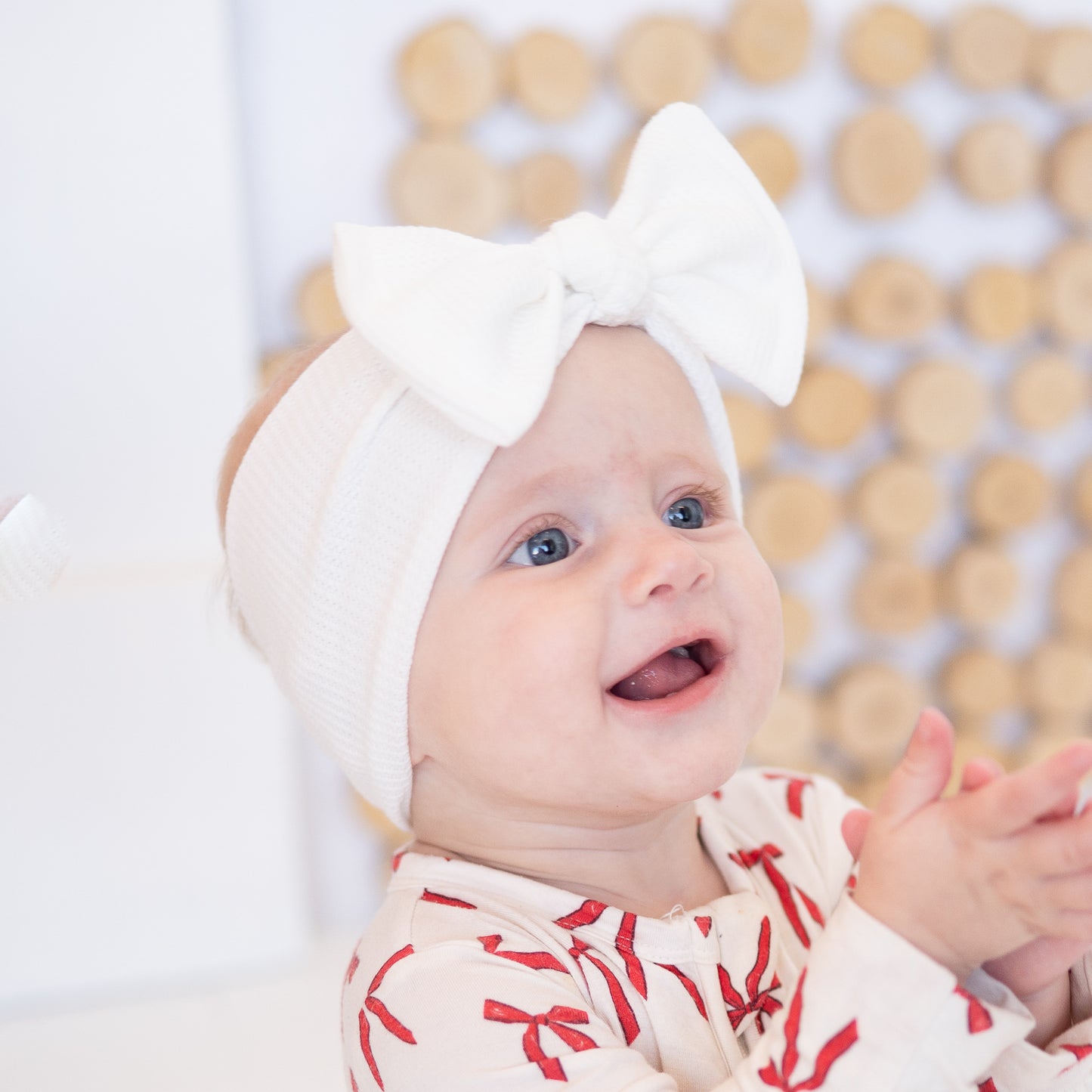 Baby wearing a white waffle top knot  headband with a large bow, sitting against a wooden wall.