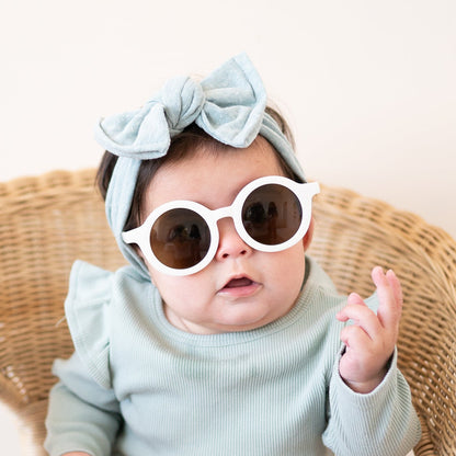 Baby wearing light blue outfit and white sunglasses with a bow headband, sitting on a wicker chair.