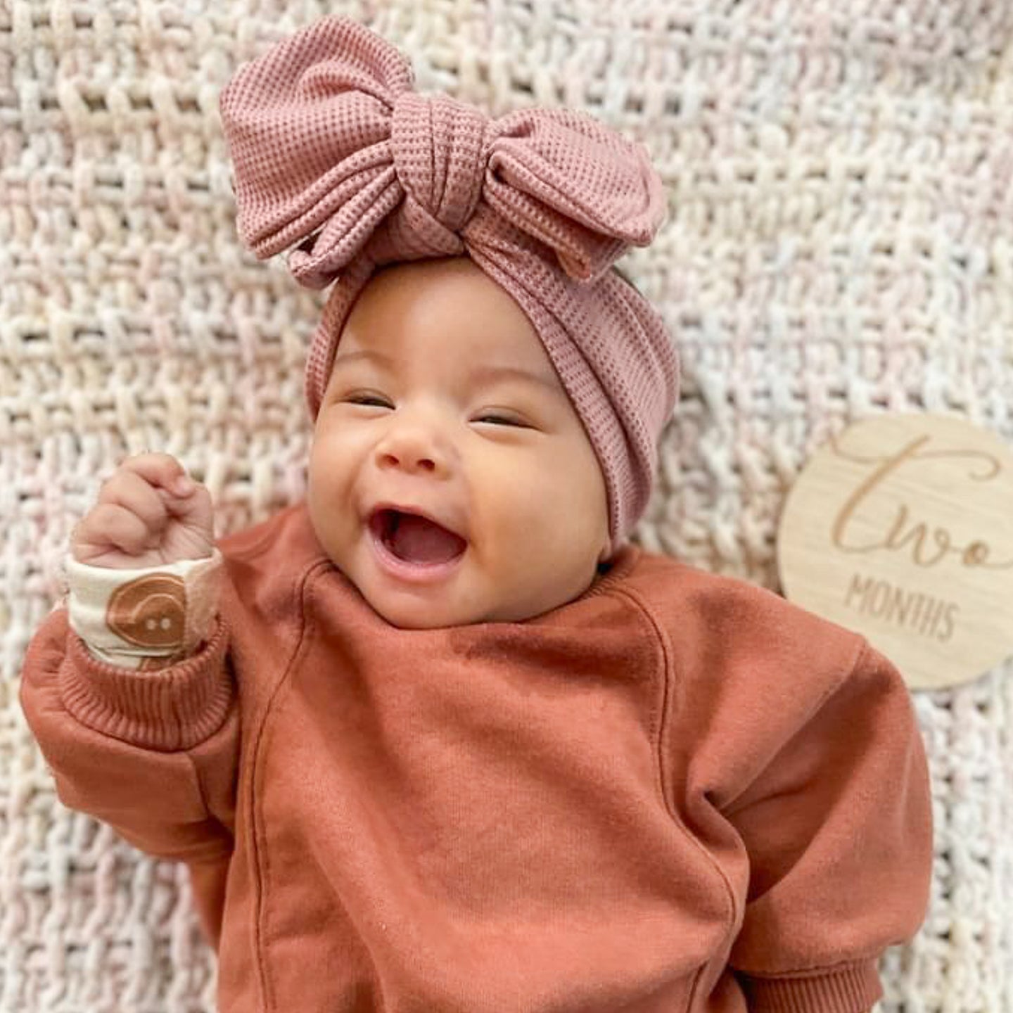 A baby wearing a mauve pink waffle headband with an oversized bow, smiling and lying on a textured surface.