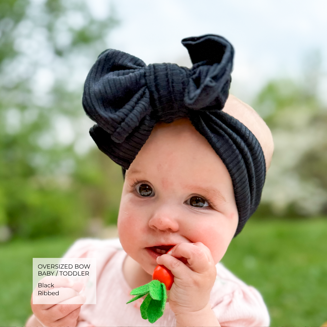 A baby wearing an oversized black ribbed bow headband, holding a carrot toy in their mouth, with a soft-focus green outdoor background. A text overlay in the bottom left reads 'Oversized Bow Baby / Toddler Black Ribbed'.