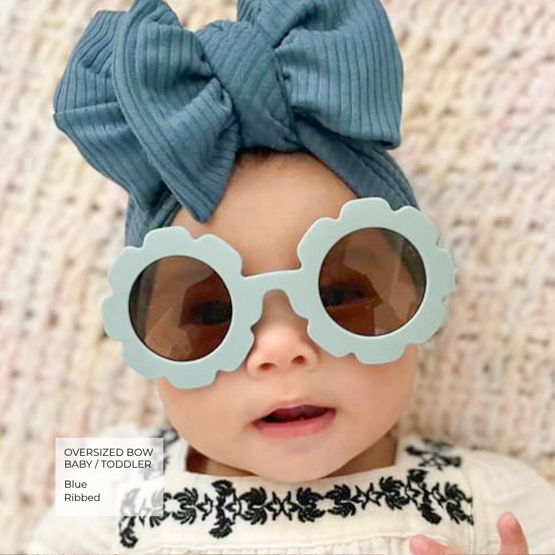 A close-up of a baby wearing oversized light blue scalloped sunglasses, a large blue ribbed bow headband, and a cream embroidered top against a soft textured beige background.