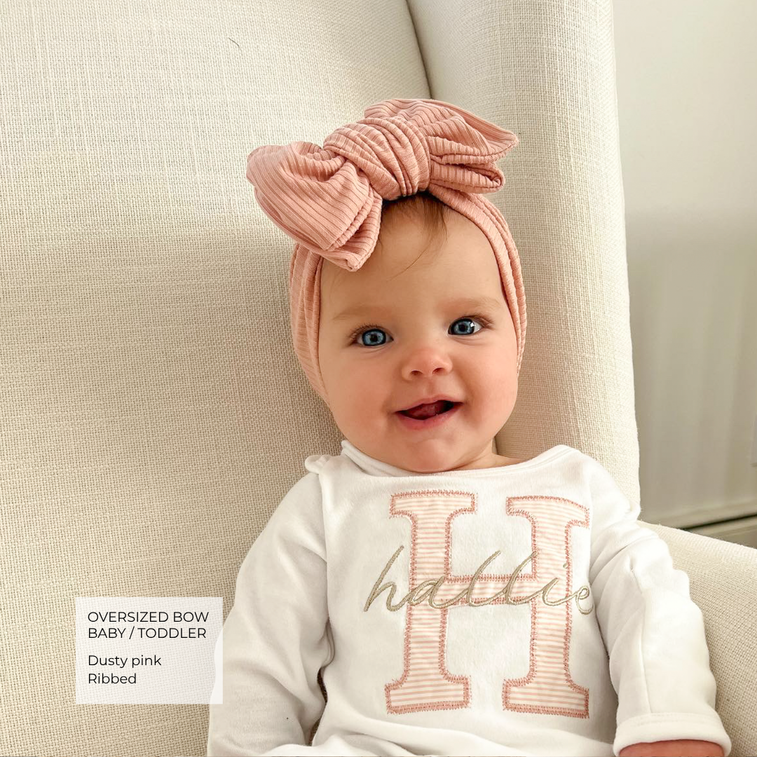 A baby wearing a dusty pink oversized bow ribbed headband and a white long-sleeve top with a large pink letter H, sitting in a beige armchair. A text box in the bottom left reads 'Oversized bow Baby / Toddler Dusty pink Ribbed'.