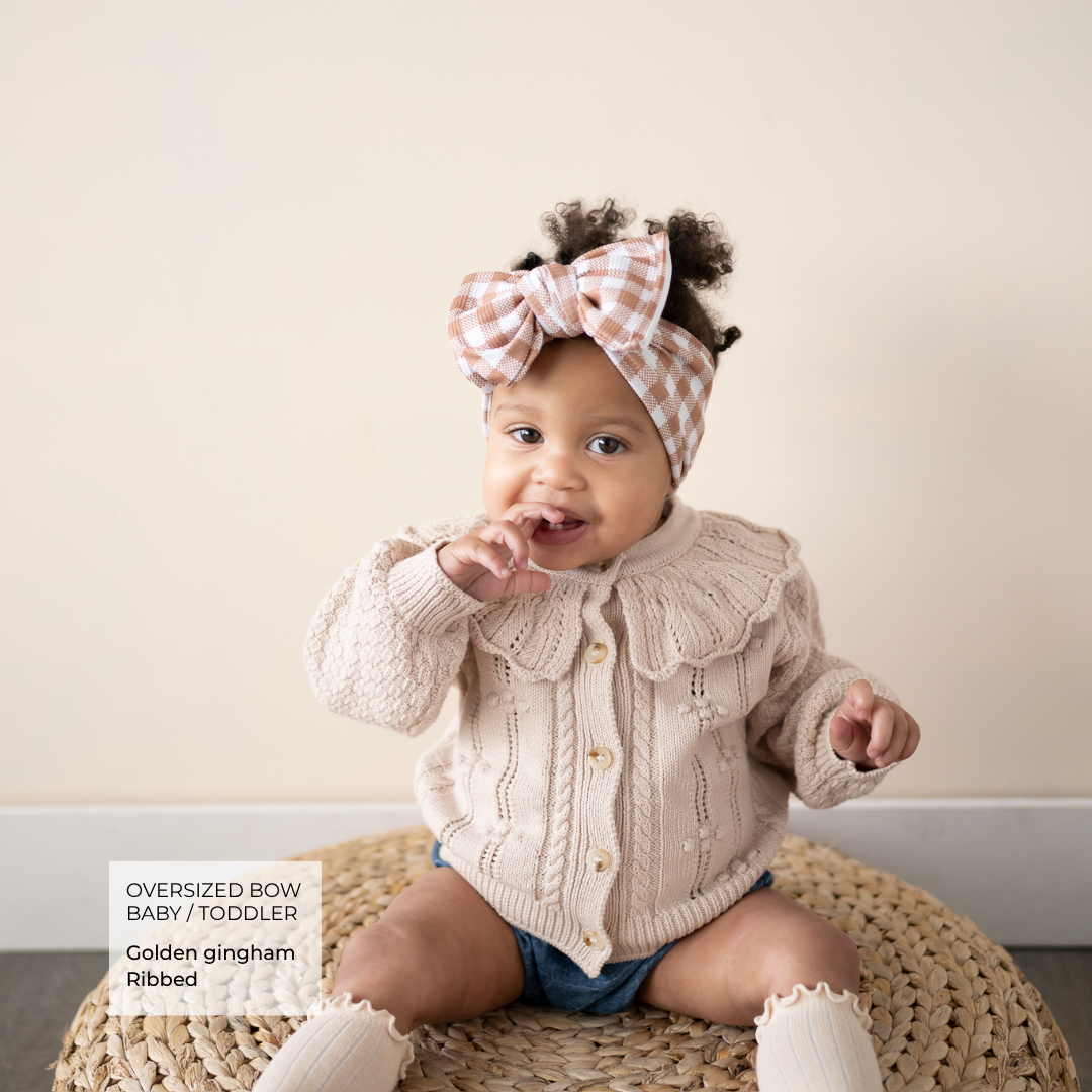 A baby wearing a cream ribbed cardigan, matching cream booties, and a pink-and-white gingham bow headband sits on a woven round rug against a beige wall. A text box in the lower left reads 'Oversized Bow Baby / Toddler Golden gingham Ribbed'.