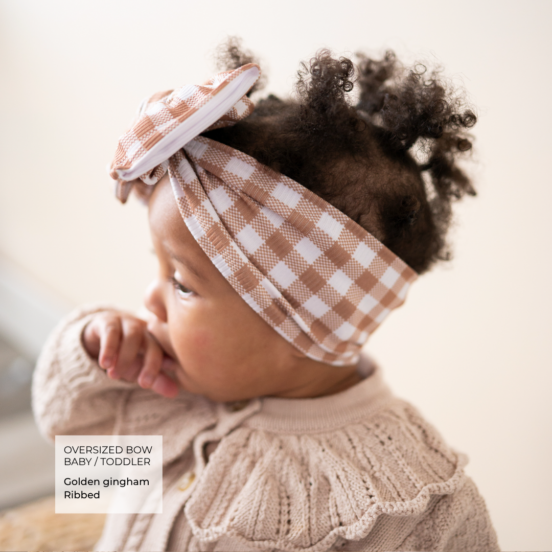 A baby wearing a beige and white gingham oversized bow headband and a matching ribbed knit sweater, shown in a warm, soft-lit portrait. Text overlay in the lower left reads 'OVERSIZED BOW BABY/TODDLER Golden gingham Golden ribbed'.