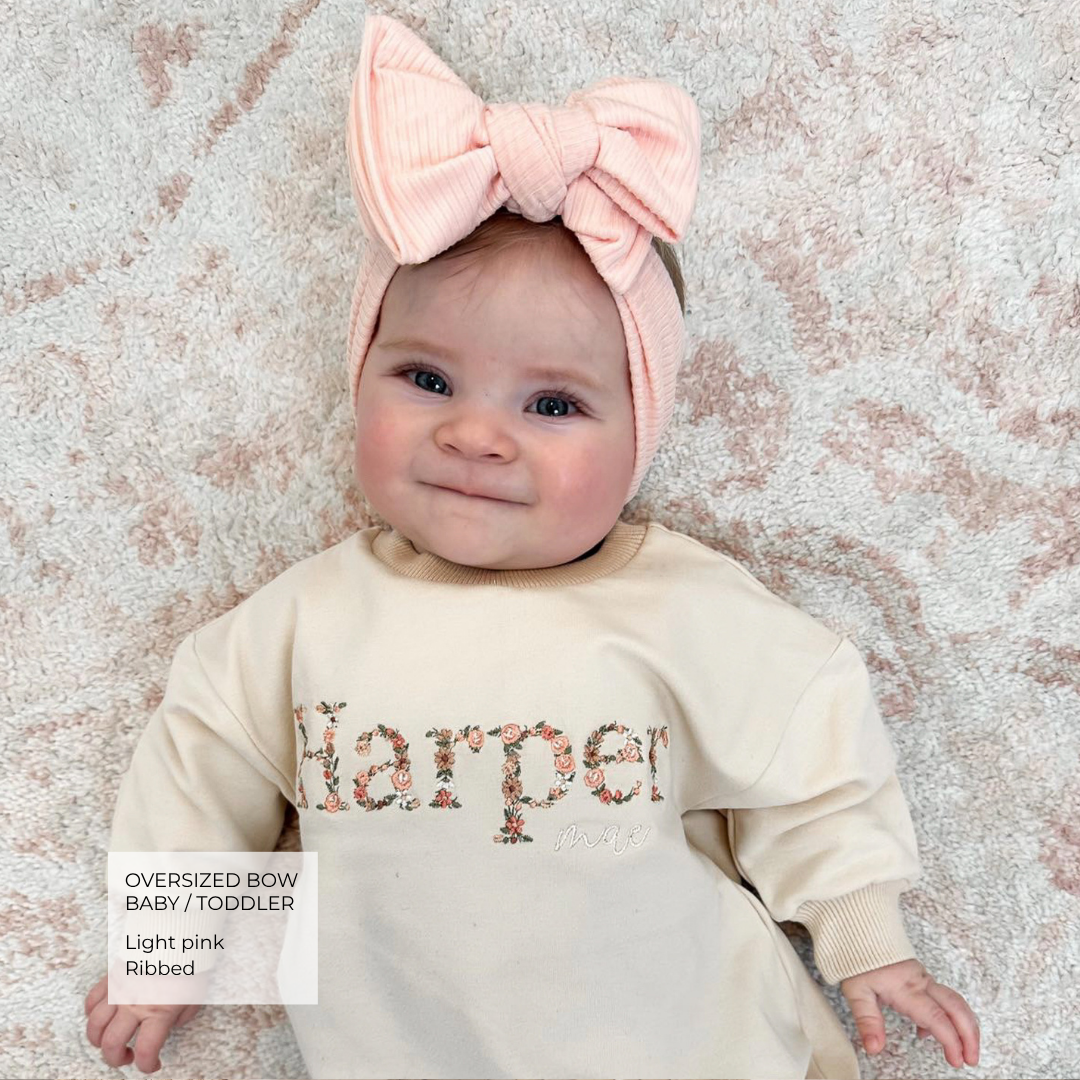 A baby lying on a textured pink and white rug, wearing a light pink oversized ribbed bow headband and a cream sweatshirt with 'Harper' in a floral patterned font. A text box in the bottom left highlights the headband's features.