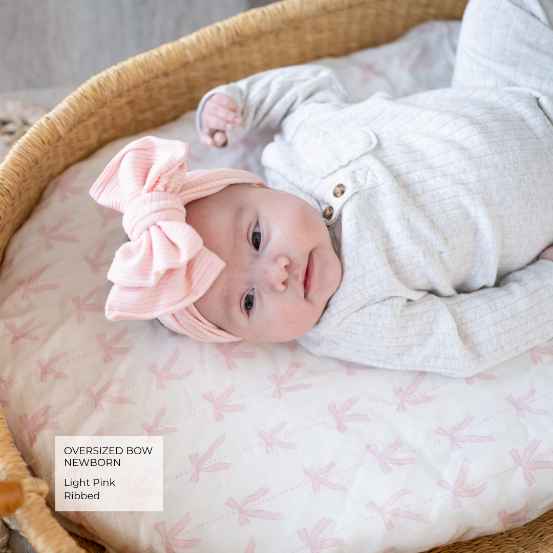 A newborn baby lying in a woven bassinet, wearing a light pink oversized bow headband and a white long-sleeve bodysuit. The bassinet cushion has a pale pink dragonfly pattern.