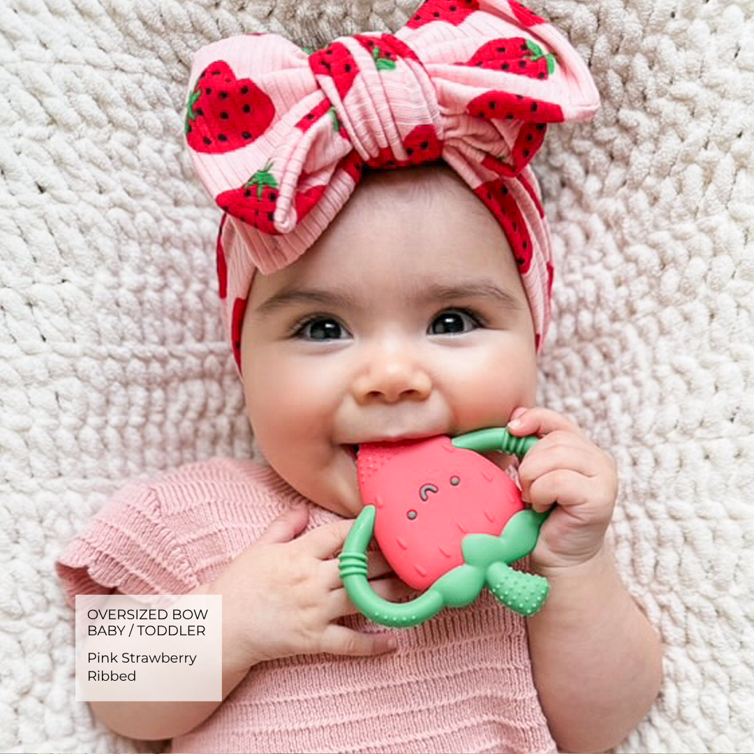 A baby lying on a textured cream blanket, wearing an oversized pink strawberry bow headband and a pink ribbed outfit, while holding a pink strawberry-shaped teether with green handles.