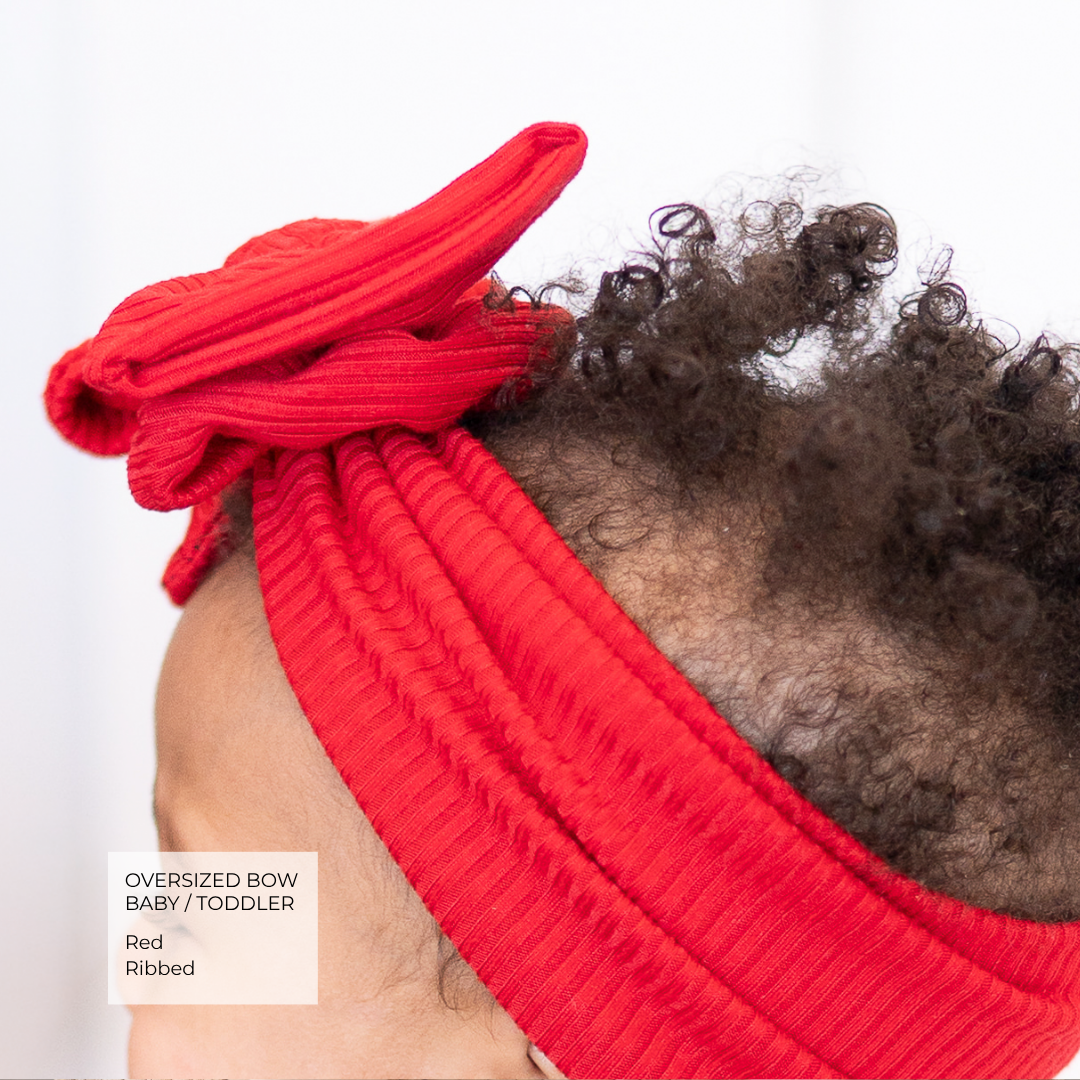A close-up of a toddler wearing a bright red ribbed knit headband with a large oversized bow, shown from the side against a white background. A text label in the lower-left corner reads 'OVERSIZED BOW BABY / TODDLER Red Ribbed'.