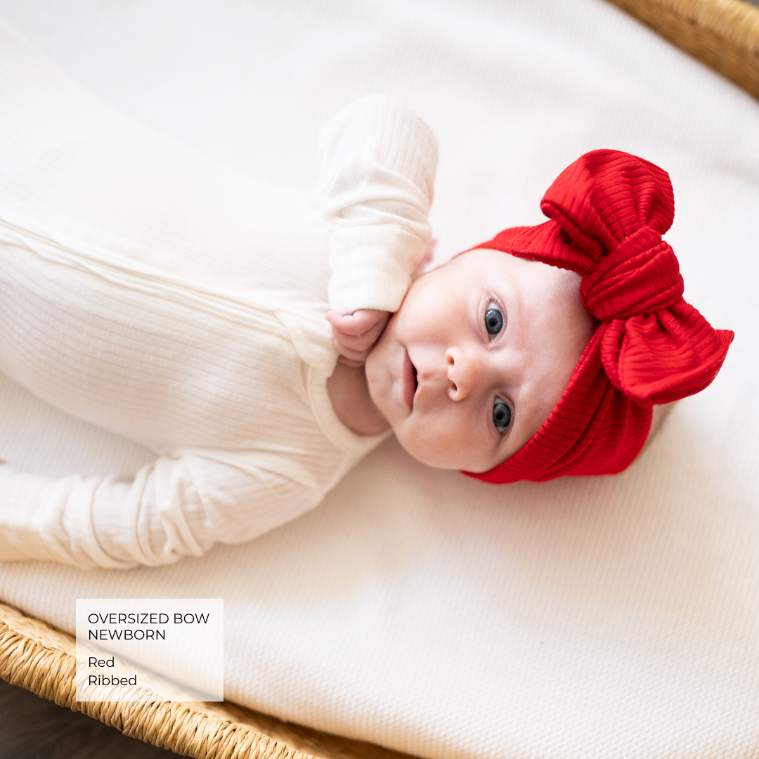 A newborn baby wearing a cream ribbed long-sleeve bodysuit and a bright red oversized bow headband, lying on a cream blanket inside a woven basket. A text box in the lower left reads 'Oversized Bow Newborn Red Ribbed'.