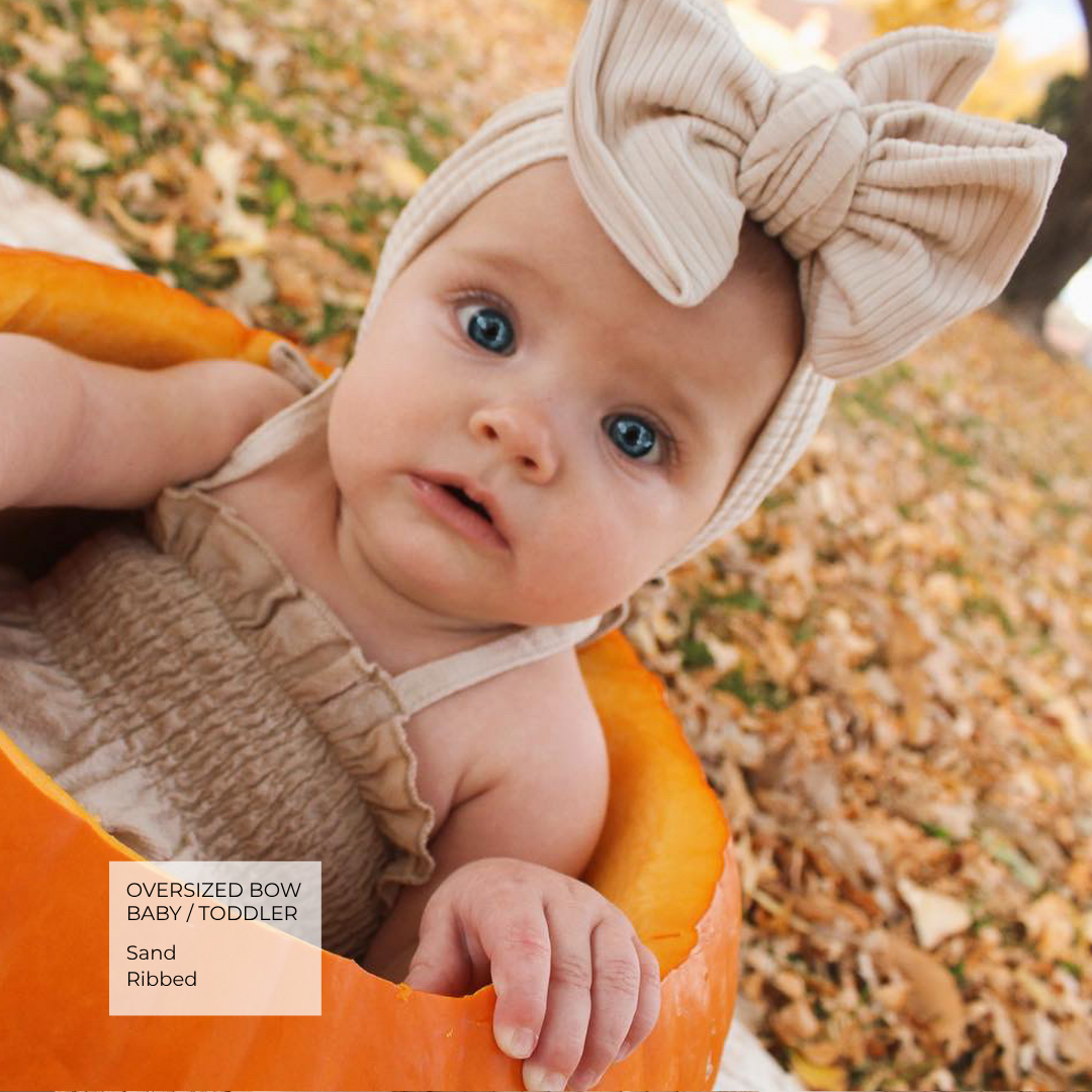 A baby wearing an oversized ribbed beige bow headband and matching ruffled bodysuit, posed inside a carved pumpkin outdoors on a leaf-covered ground. Text overlay reads 'Oversized Bow Baby / Toddler Sand Ribbed'.
