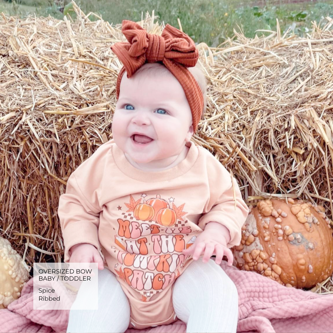 A baby sitting outdoors in front of a hay bale and pumpkins, wearing a peach long-sleeve bodysuit with a pumpkin graphic and a rust-colored oversized bow headband. A text box in the lower left reads 'OVERSIZED BOW BABY/TRODDER Spice Ribbed'.