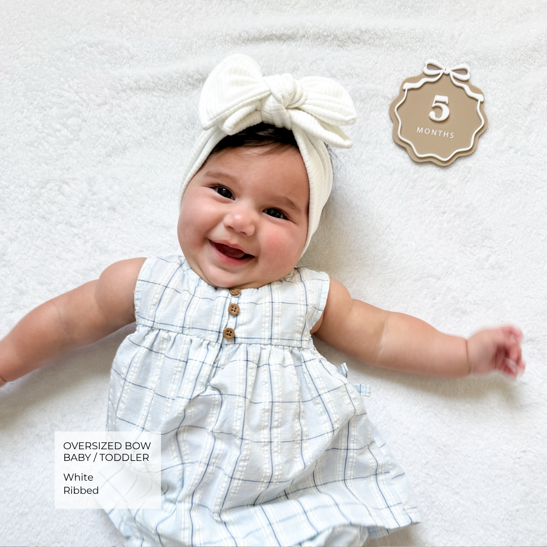 A smiling baby lying on a white textured blanket, wearing an oversized white bow headband and a white ribbed sleeveless romper with a light blue grid pattern and small gold buttons. A badge in the top right says 5 months, and a text box in the bottom left highlights the oversized bow, baby/toddler fit, and white ribbed fabric.