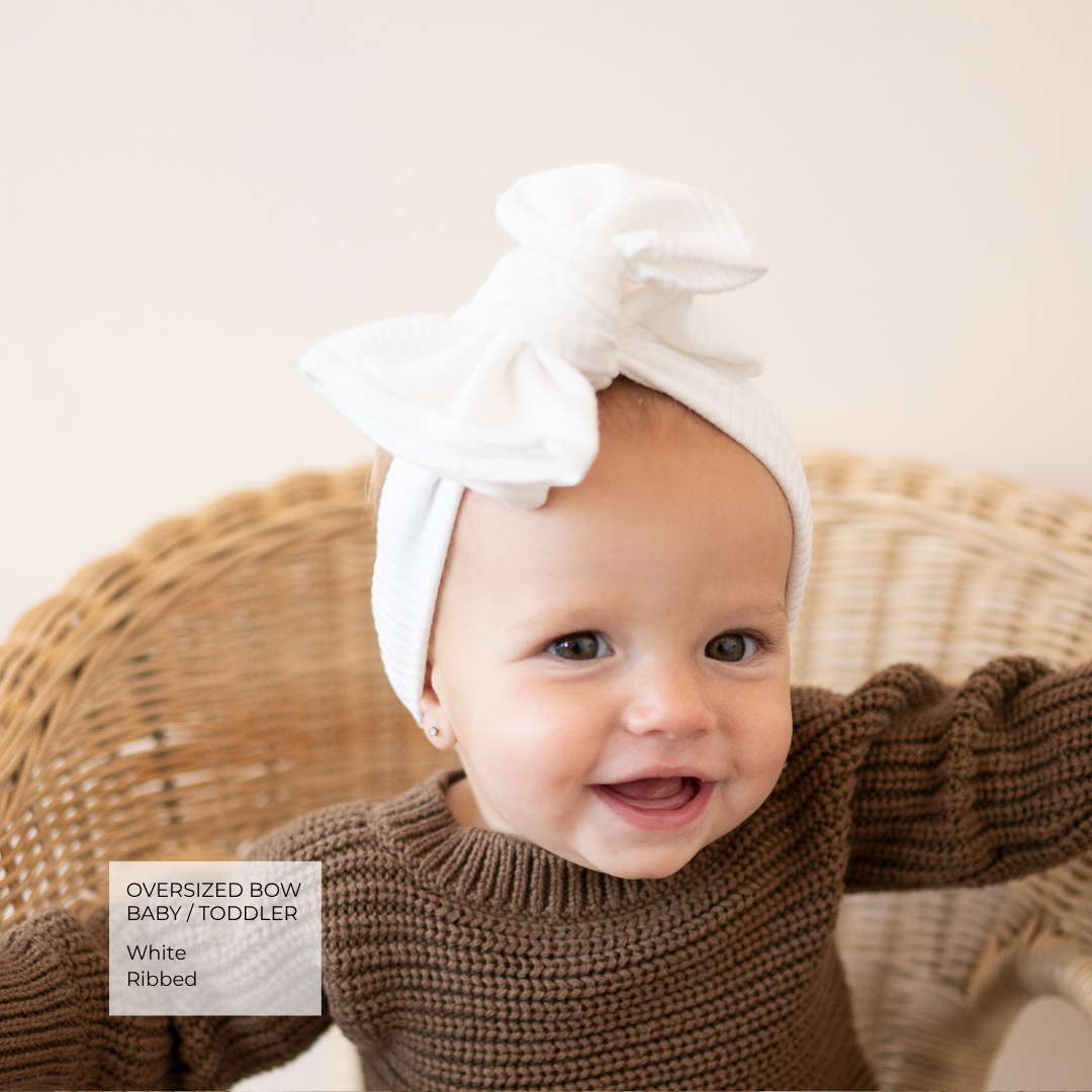 A smiling baby wearing an oversized white ribbed bow headband and a brown knit sweater, seated in a wicker chair against a soft cream background. A text box in the lower left reads 'Oversized Bow Baby / Toddler White Ribbed'.