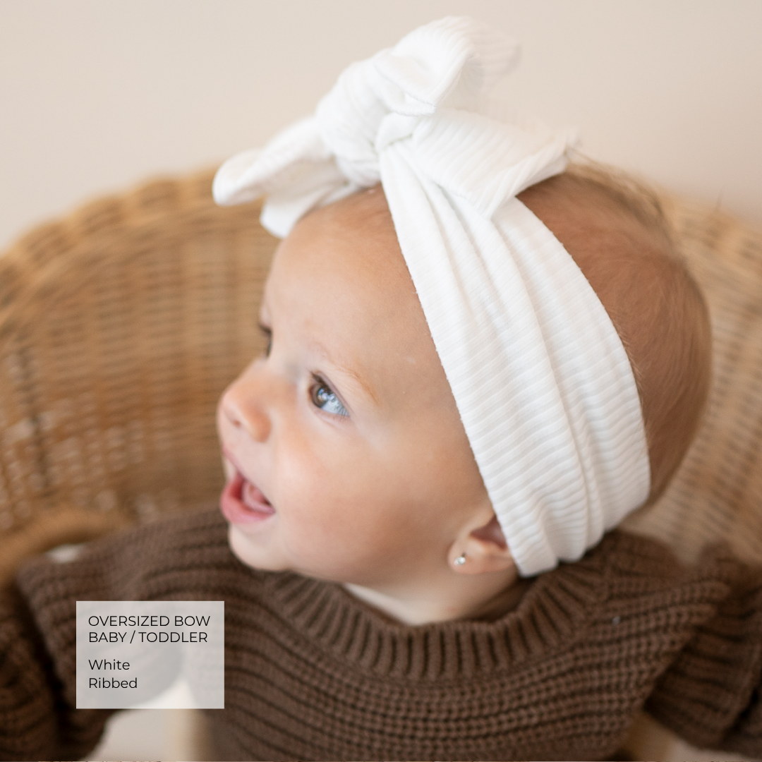 A baby wearing a white ribbed oversized bow headband, sitting in a wicker basket. A text overlay in the bottom left corner reads 'Oversized Bow Baby / Toddler White Ribbed'.