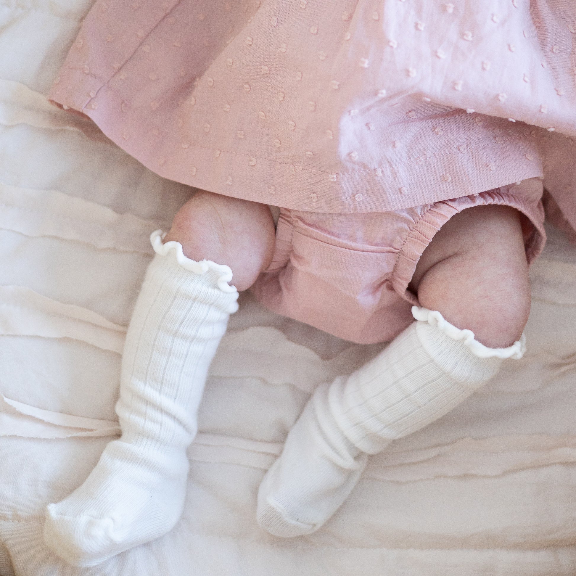 Baby legs wearing white knee socks and pink outfit on a soft surface