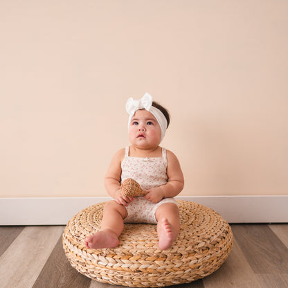 A baby sitting on a woven mat wearing a white eyelet headband and a white outfit.