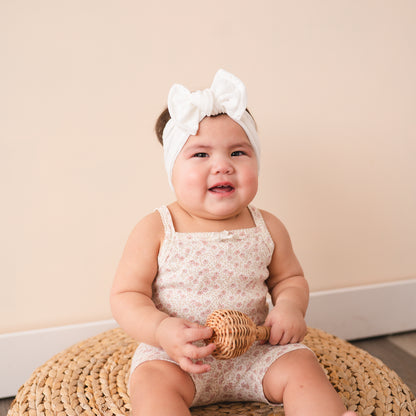 Baby sitting on woven pouf smiling and wearing a white eyelet headband.