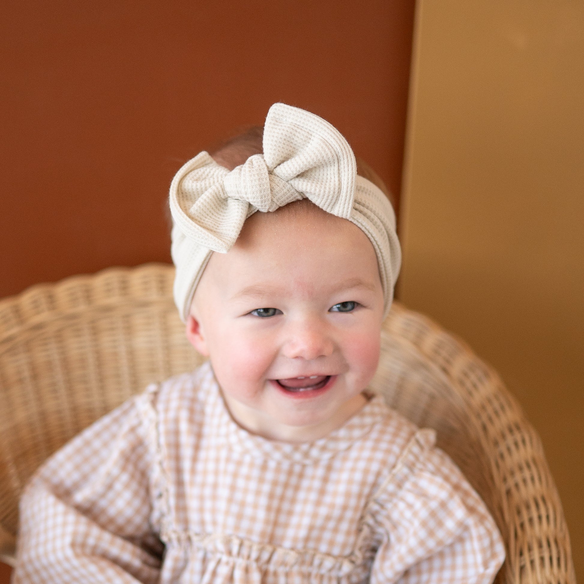 Baby wearing a beige waffle textured headband with a large top knot bow, sitting on a wicker chair.