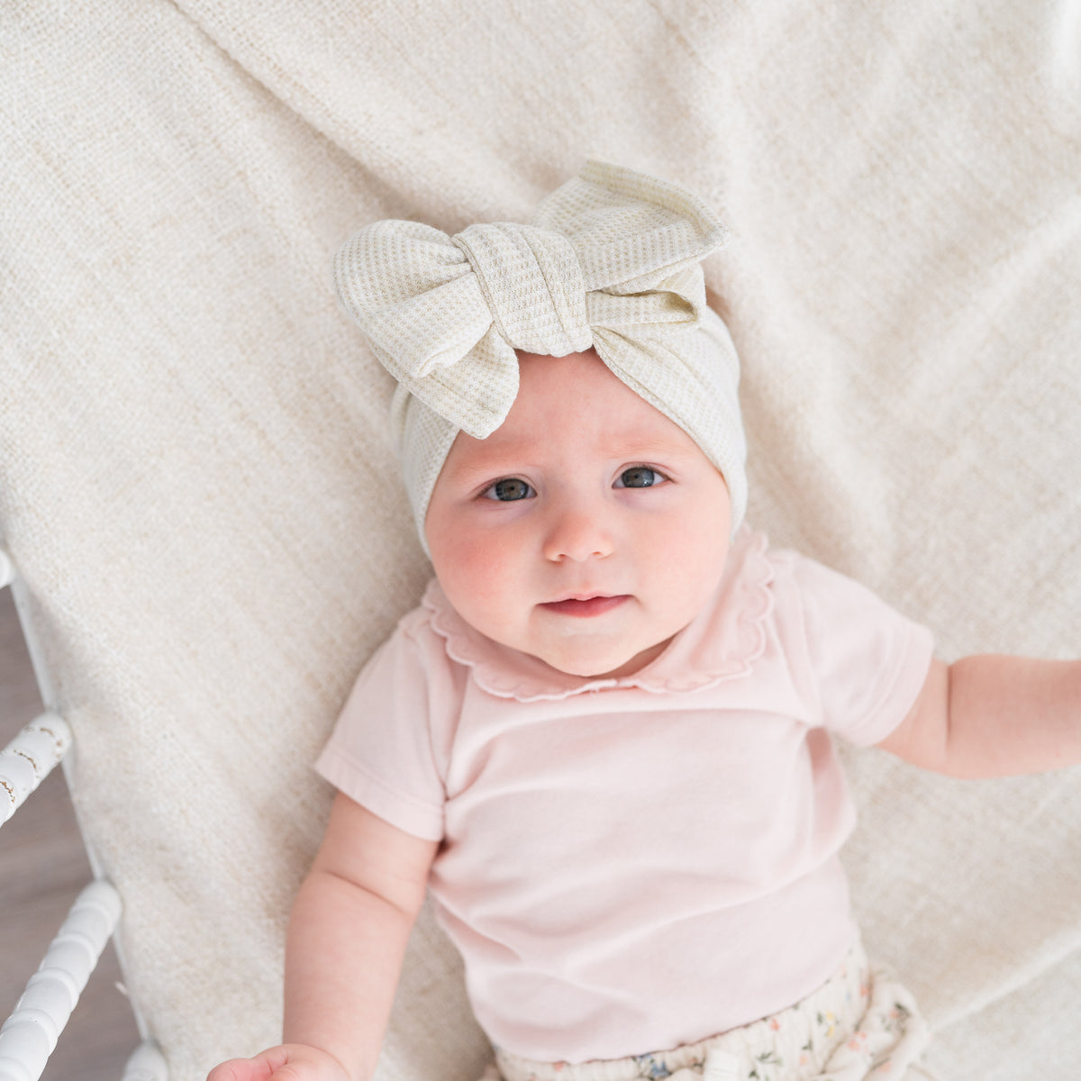 Baby wearing a beige waffle knit headband with a bow, sitting on a soft surface.