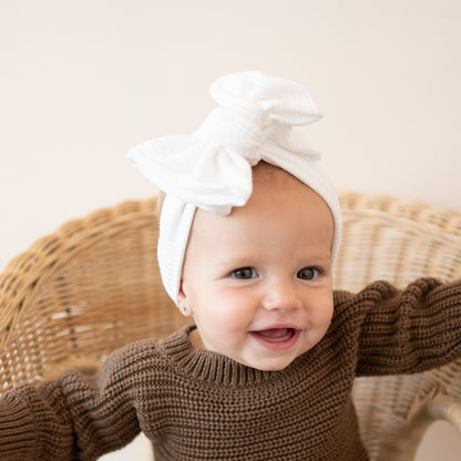 Baby wearing a brown sweater and white ribbed oversized bow headband, sitting in a wicker chair.