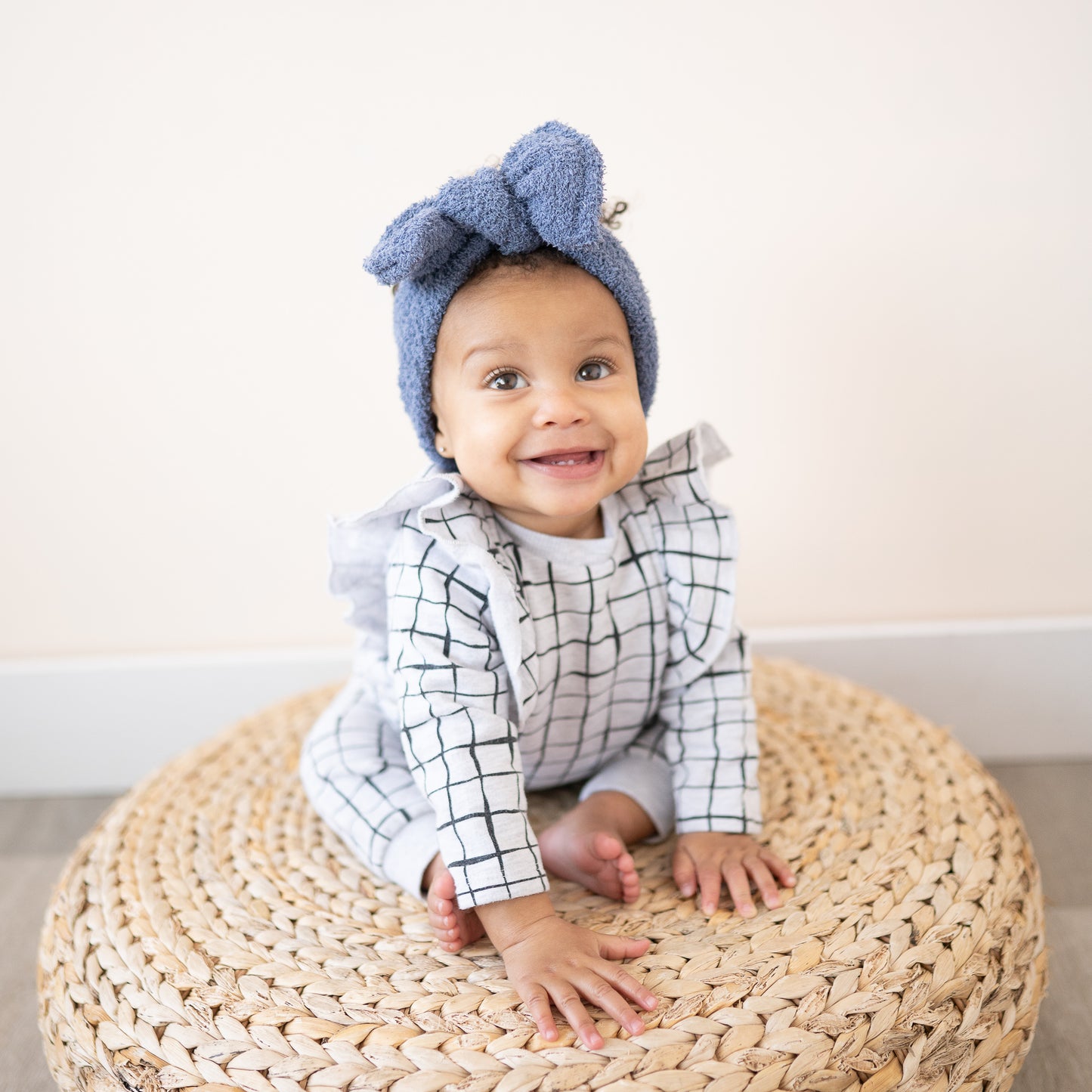 Baby wearing a blue fuzzy headband bow and checkered outfit sitting on a woven stool.
