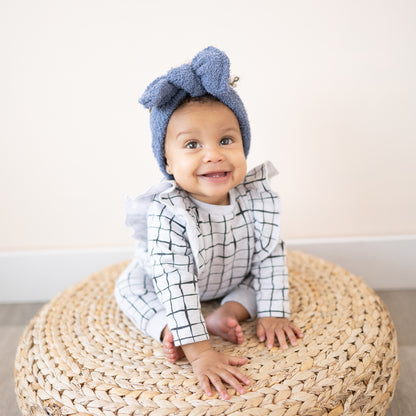 Baby wearing a blue fuzzy headband bow and checkered outfit sitting on a woven stool.
