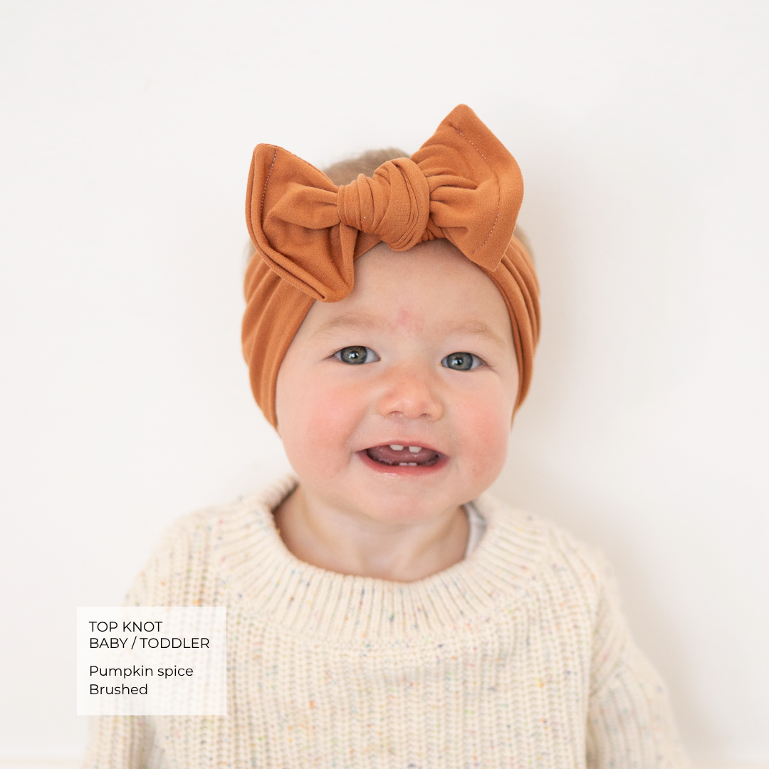 A baby wearing a pumpkin spice brown velvet top knot headband and a cream knit sweater, photographed against a clean white background with a text overlay in the bottom left.